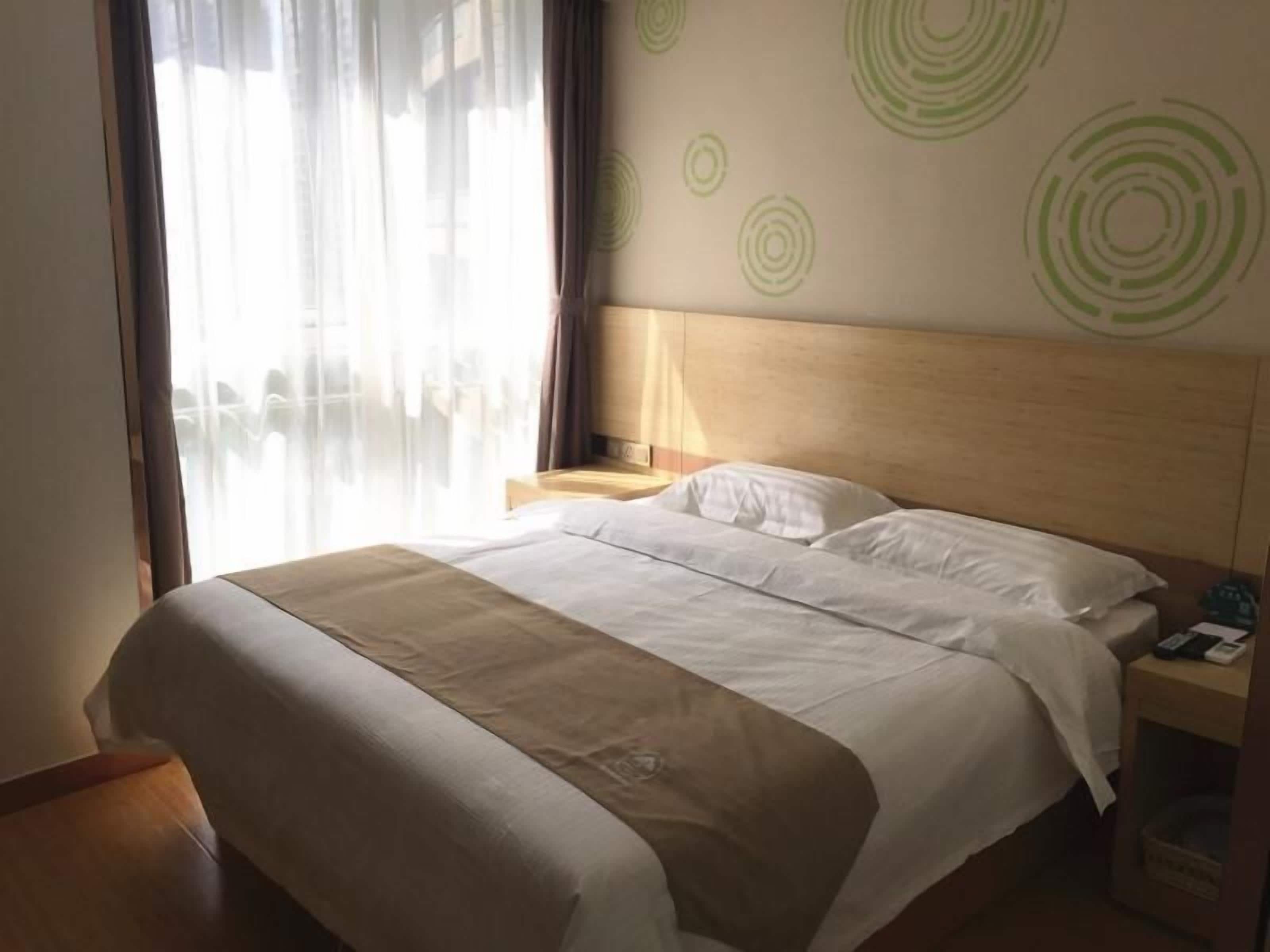 A hotel room with a large bed, a wooden headboard, a nightstand with a lamp, and a view of the city through sheer curtains.