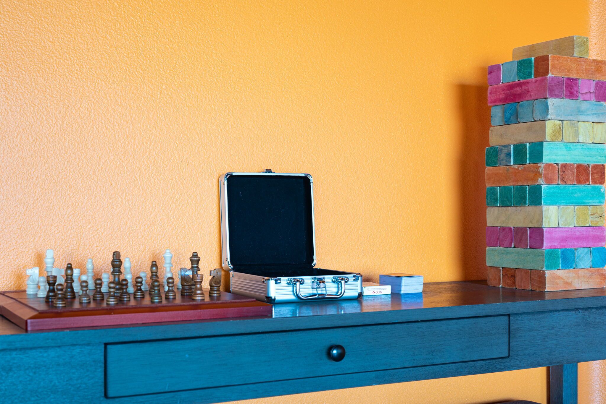 A chessboard, a suitcase, and a stack of colorful blocks on a desk against an orange wall.