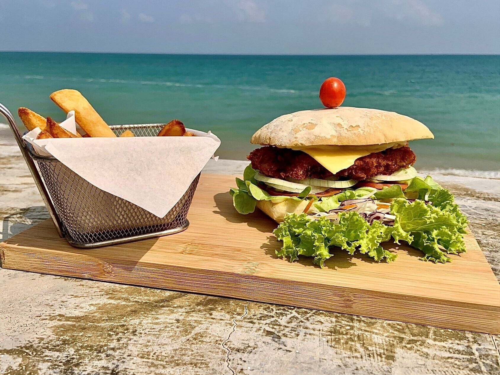 A burger with a tomato on top, fries in a basket, and lettuce on a wooden board by the sea.