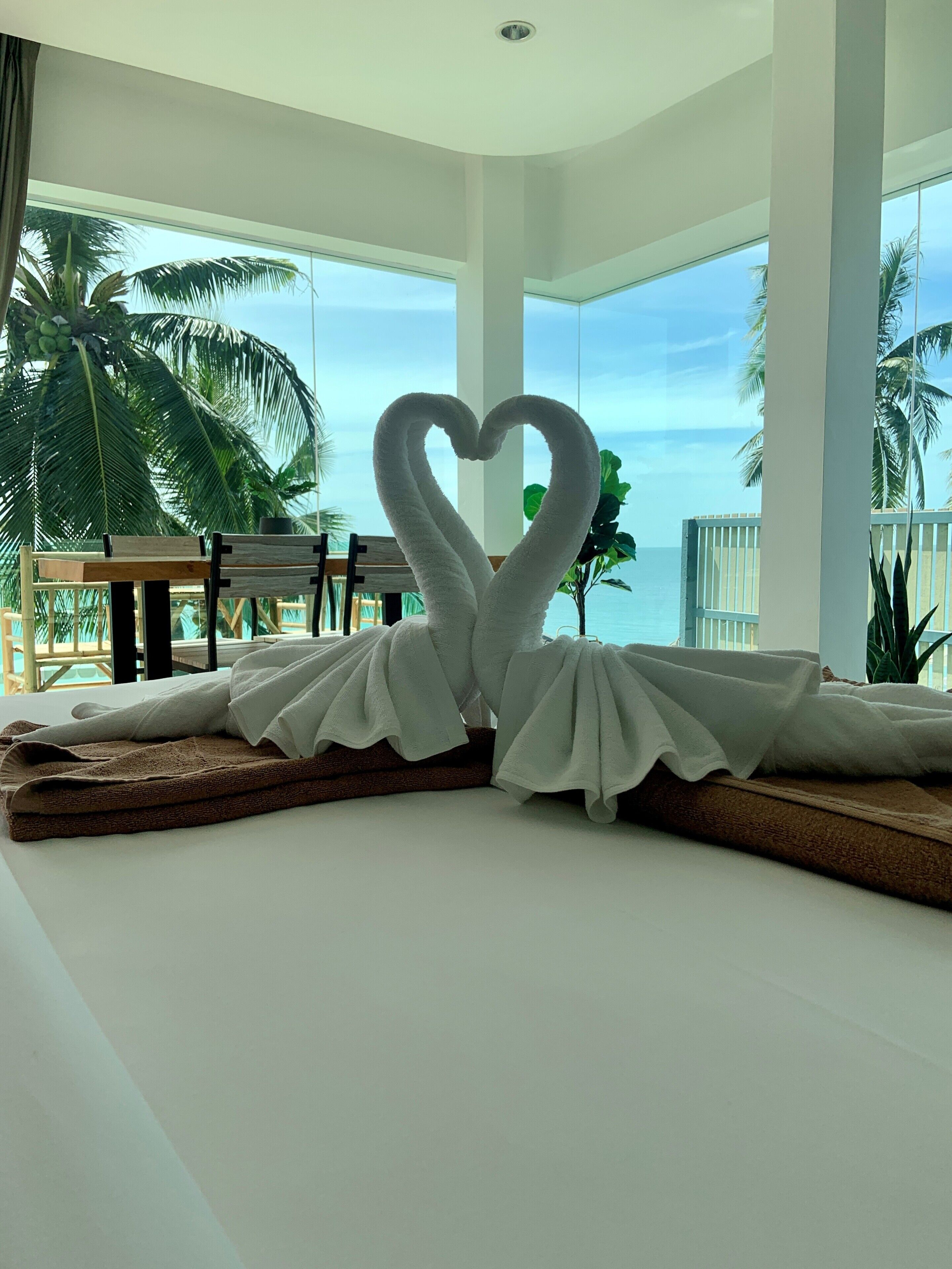 Una habitación con una toalla dispuesta en forma de corazón sobre la cama, ventanas amplias y vistas a palmeras y al océano.