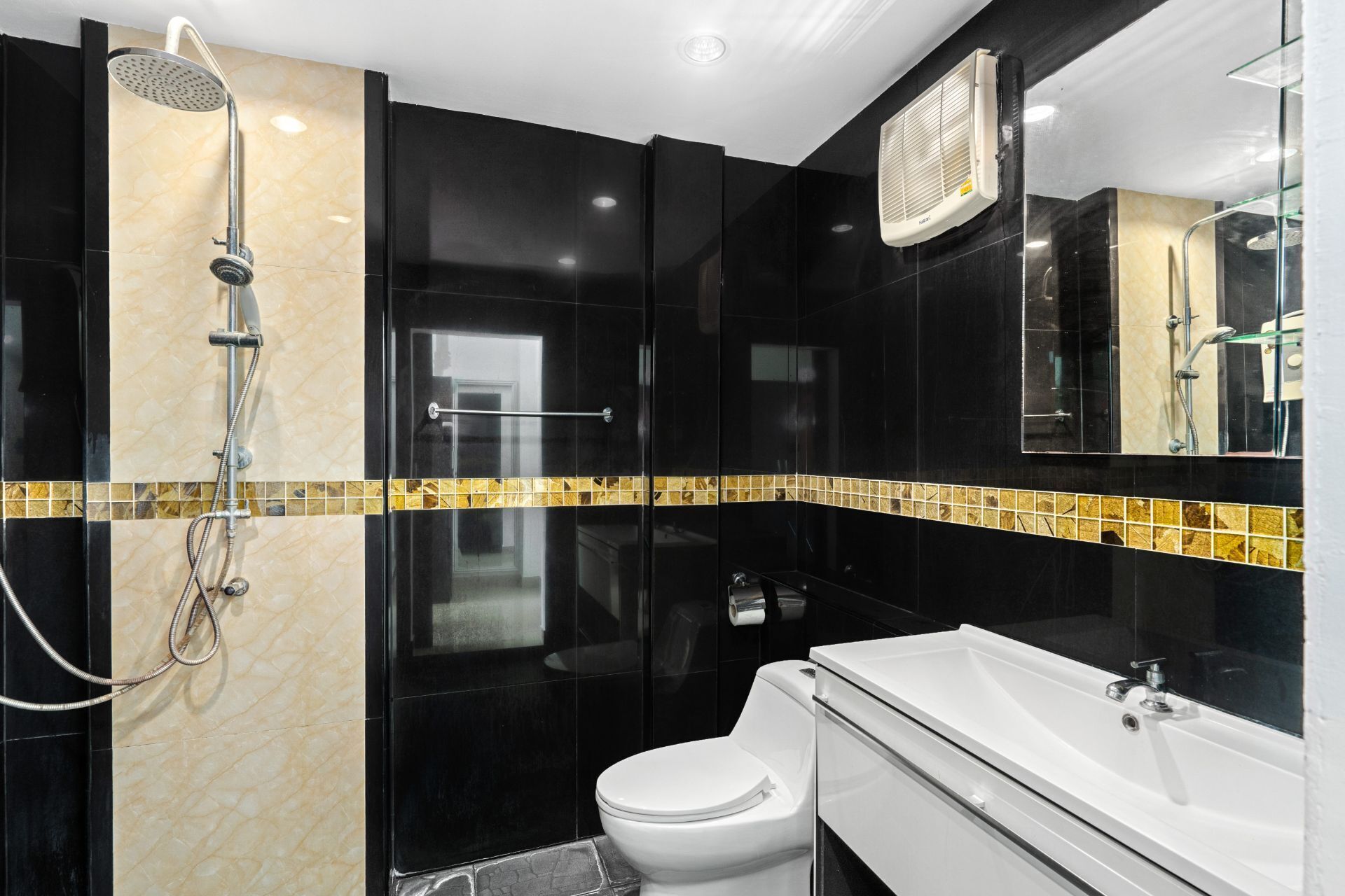 Une salle de bain moderne avec un mur recouvert de carreaux noirs et beiges, des toilettes blanches, un lavabo blanc et une douche équipée d’un pommeau de douche à main.