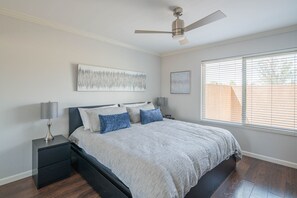 Une chambre à coucher comprenant un lit, des tables de chevet, un ventilateur de plafond, une fenêtre avec des stores et des œuvres d’art accrochées au mur.