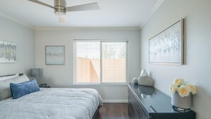 Une chambre à coucher comprenant un lit, une commode, une table de chevet, un ventilateur de plafond et une fenêtre avec des stores.