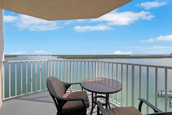 A balcony with a view of a calm body of water, a round table, and two wicker chairs.