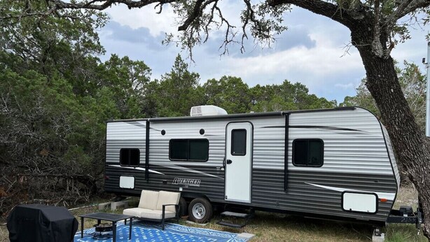 Un camping-car est garé dans une zone boisée, à proximité d’un barbecue couvert, d’un tapis à motifs bleus et d’un canapé blanc.