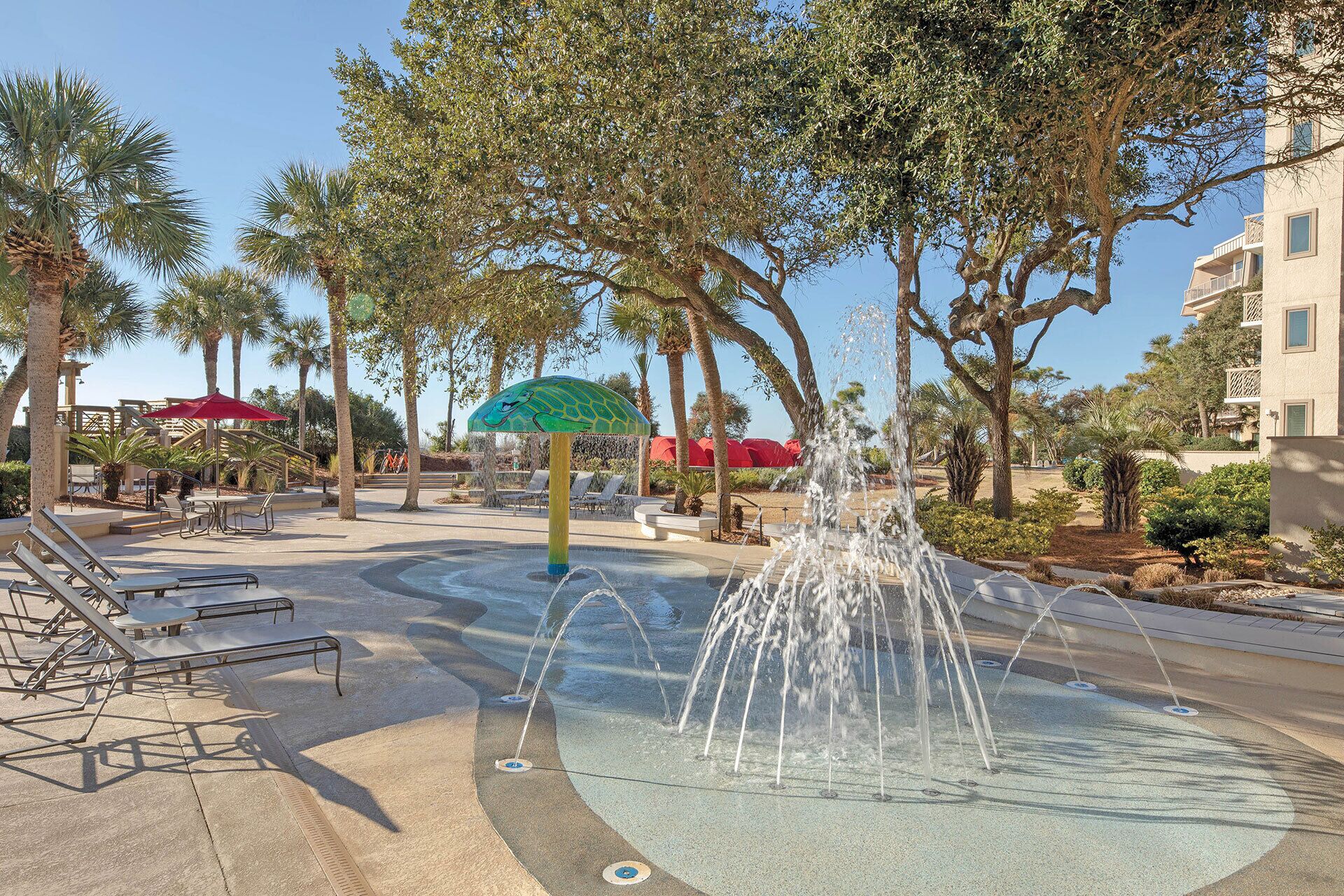 A water fountain with a green umbrella, surrounded by palm trees and lounge chairs.