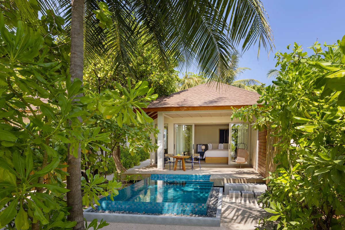 A poolside area with a view of a bedroom and a dining area, surrounded by lush greenery and palm trees.