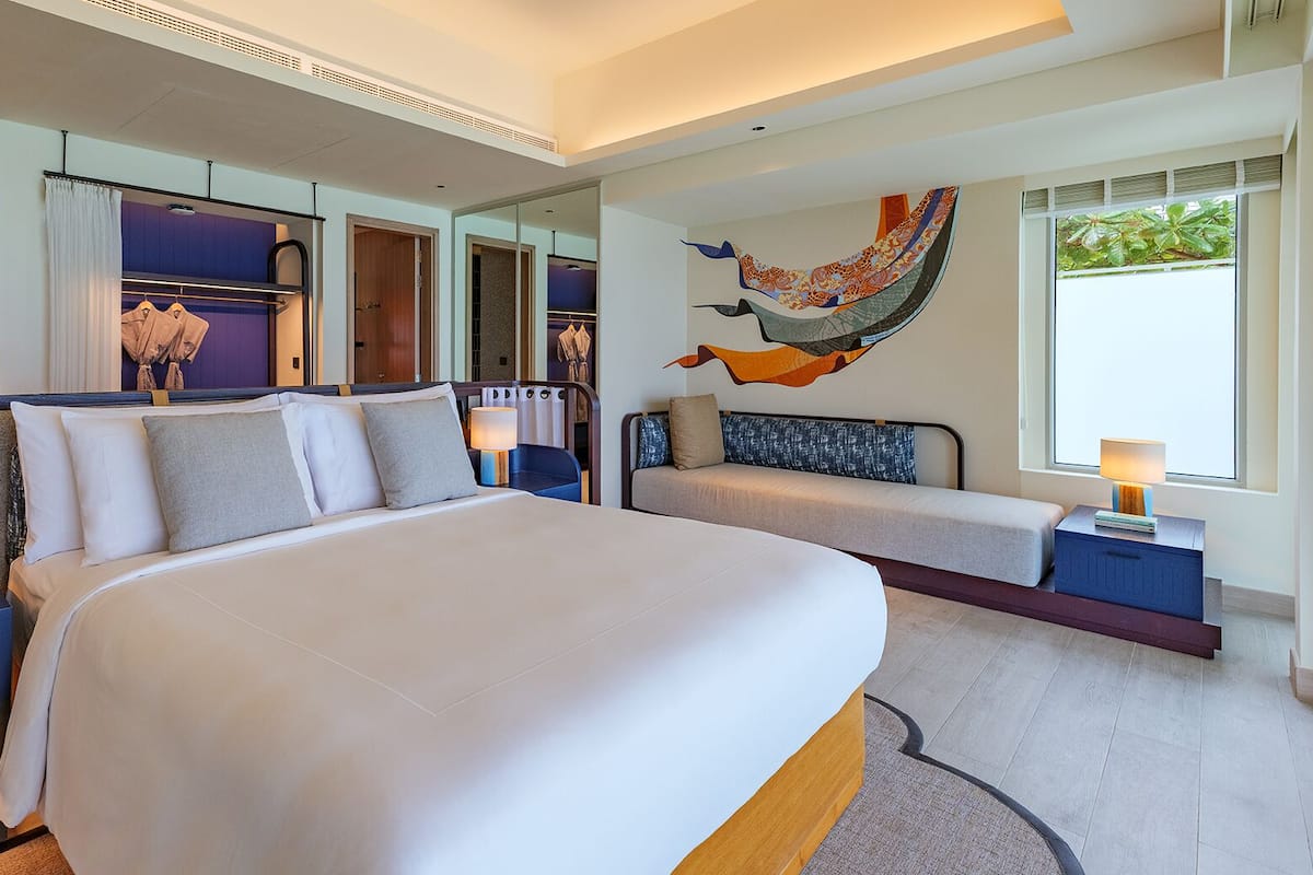 A modern hotel room with a large bed, bedside tables with lamps, a seating area with a sofa, and a large window with a view of greenery.