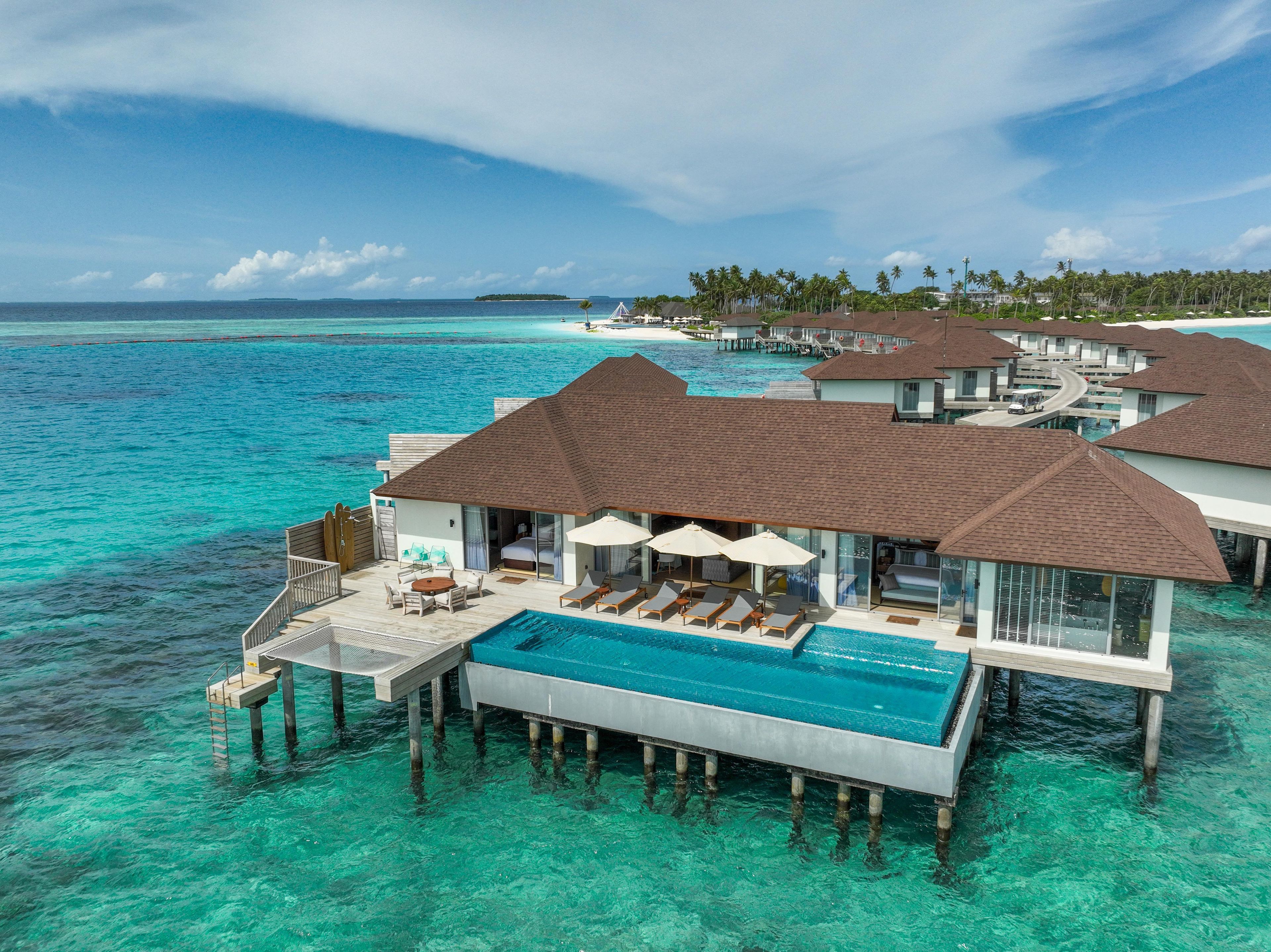 Cabañas sobre el agua con piscinas infinitas, un mar azul cristalino y una isla tropical al fondo.