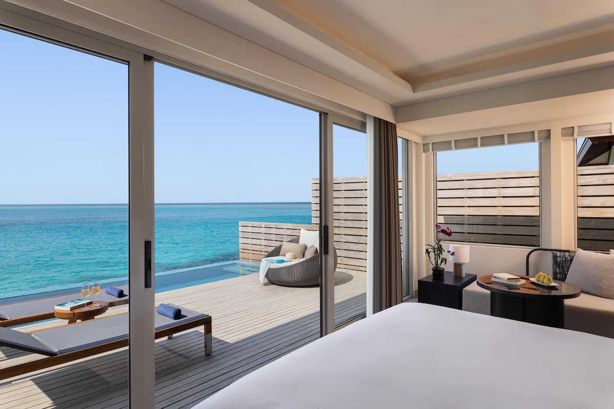 A modern hotel room with a large bed, a seating area with a table and chairs, a small side table with a lamp, and a view of the ocean.