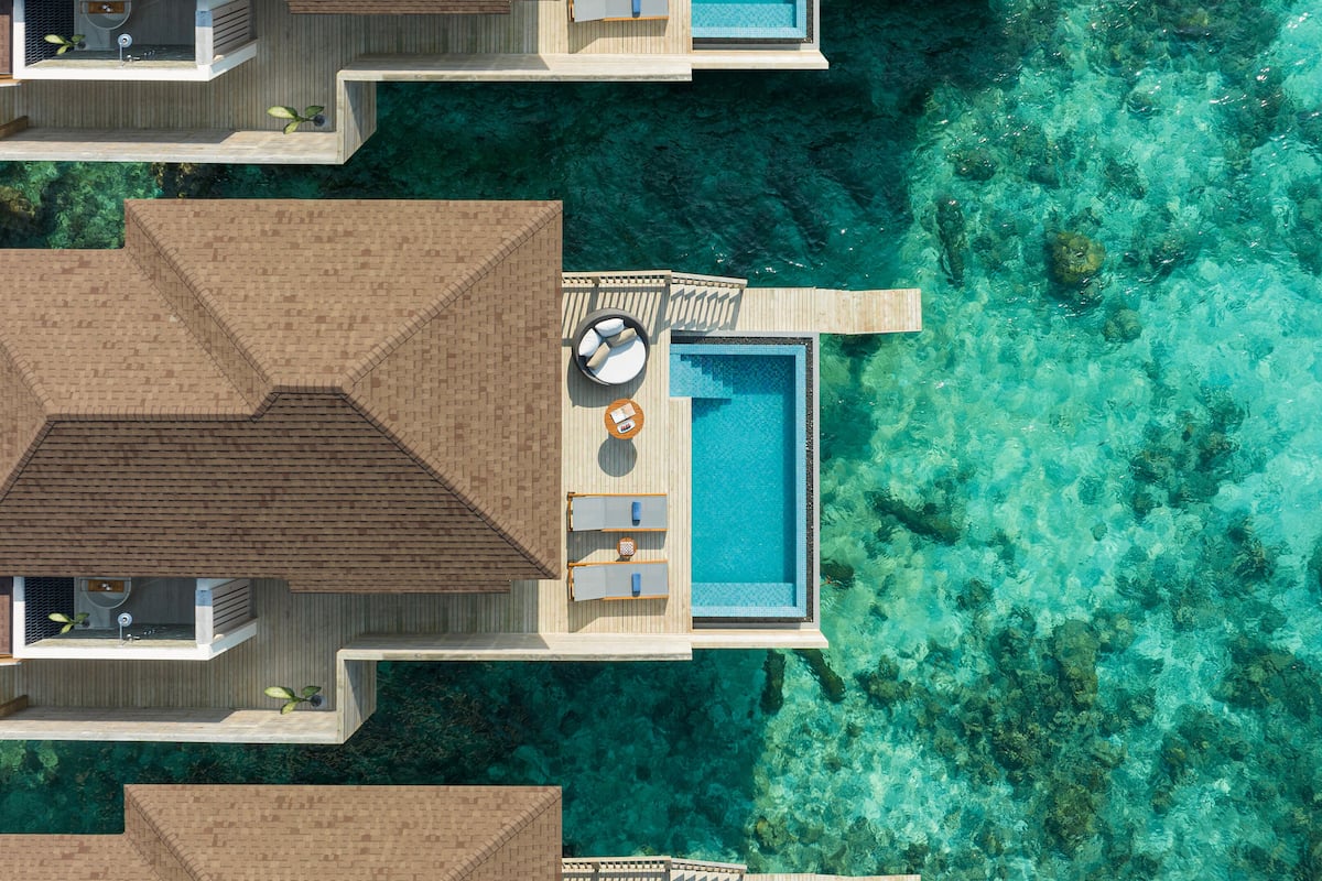An aerial view of a resort with overwater bungalows, a central pool, and clear turquoise water.