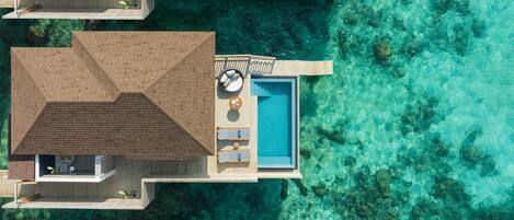 An aerial view of a resort with overwater bungalows, a central pool, and clear turquoise water.