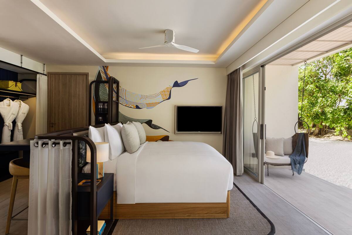 A modern hotel room with a large bed, a desk with a lamp, a television, and a view of the beach.