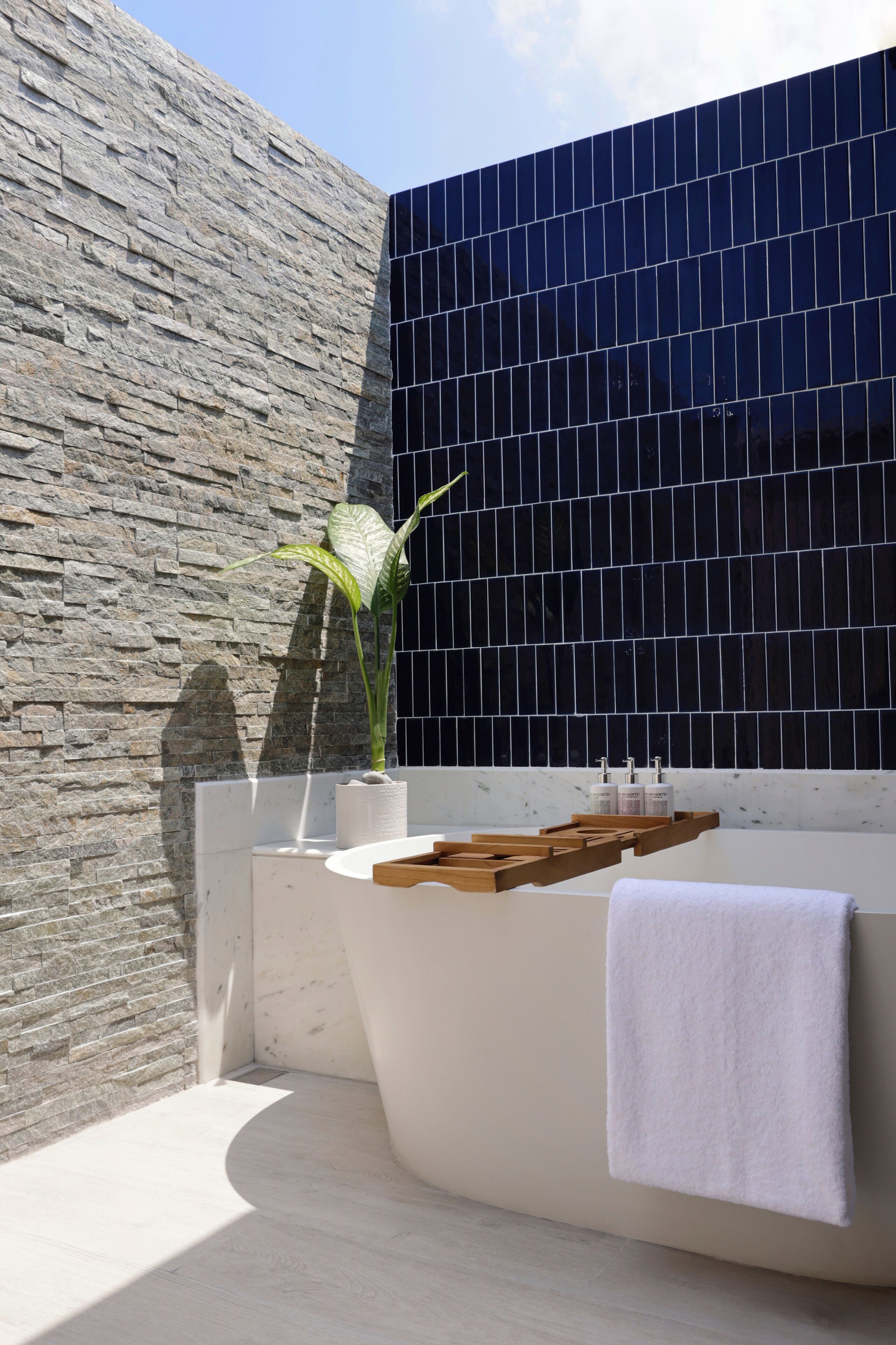 Un baño moderno con pared de piedra, pared con azulejos negros, bañera, estantes de madera y una planta en maceta.