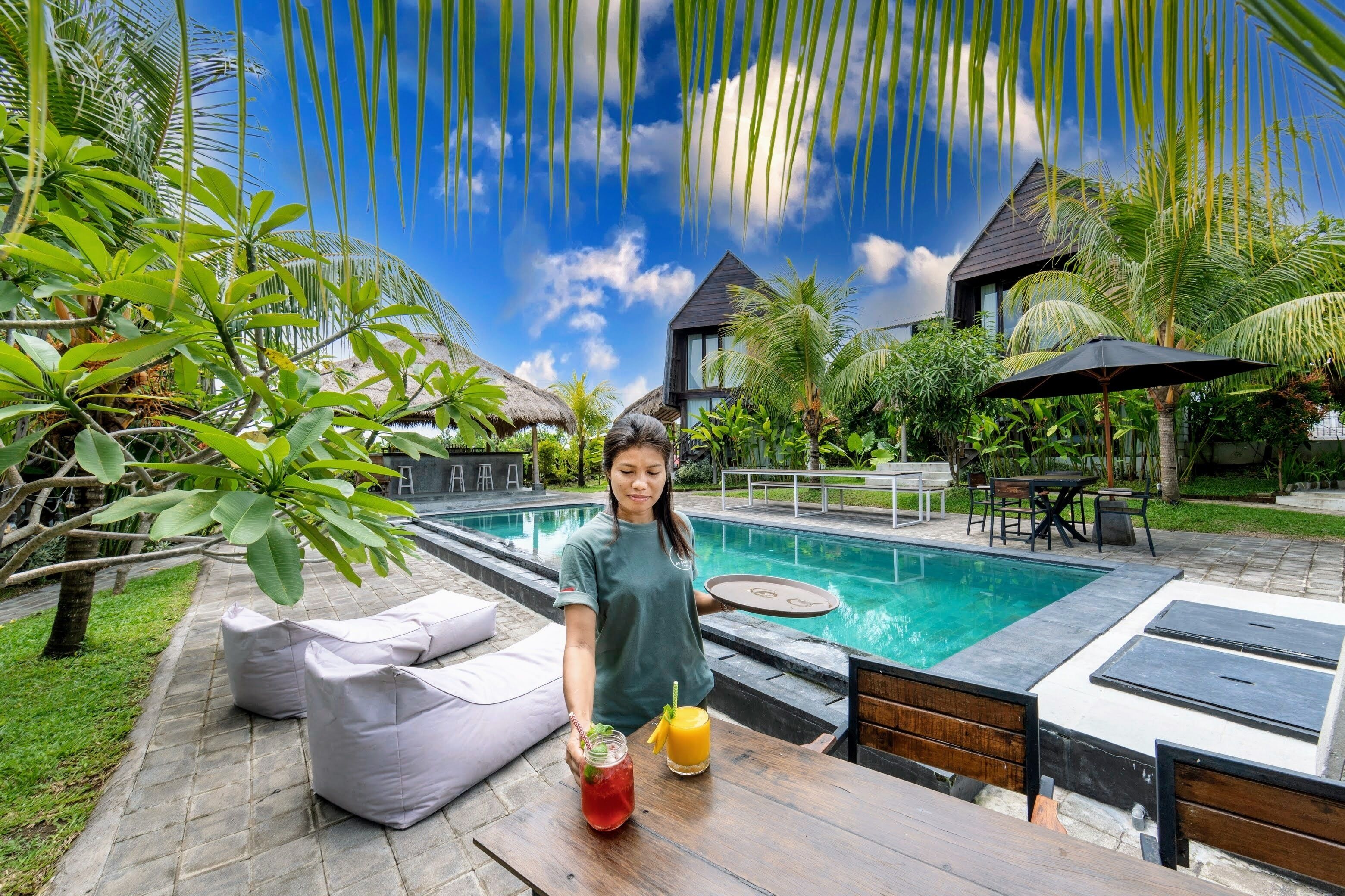 A poolside area with a table, cushions, and a person holding a drink.