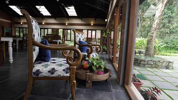 A covered outdoor seating area with wooden chairs and tables, surrounded by large windows and potted plants. - Magnolia Estates & Resort (Virajpet)