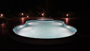 A circular swimming pool with a tiled edge and a central fountain, surrounded by a dark pool deck and illuminated by multiple lights. - Magnolia Estates & Resort (Virajpet)