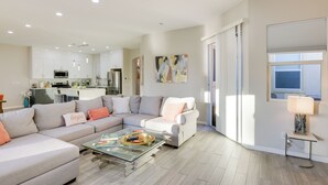 Una sala de estar moderna con un sofá seccional, una mesa de centro de vidrio y una cocina al fondo.