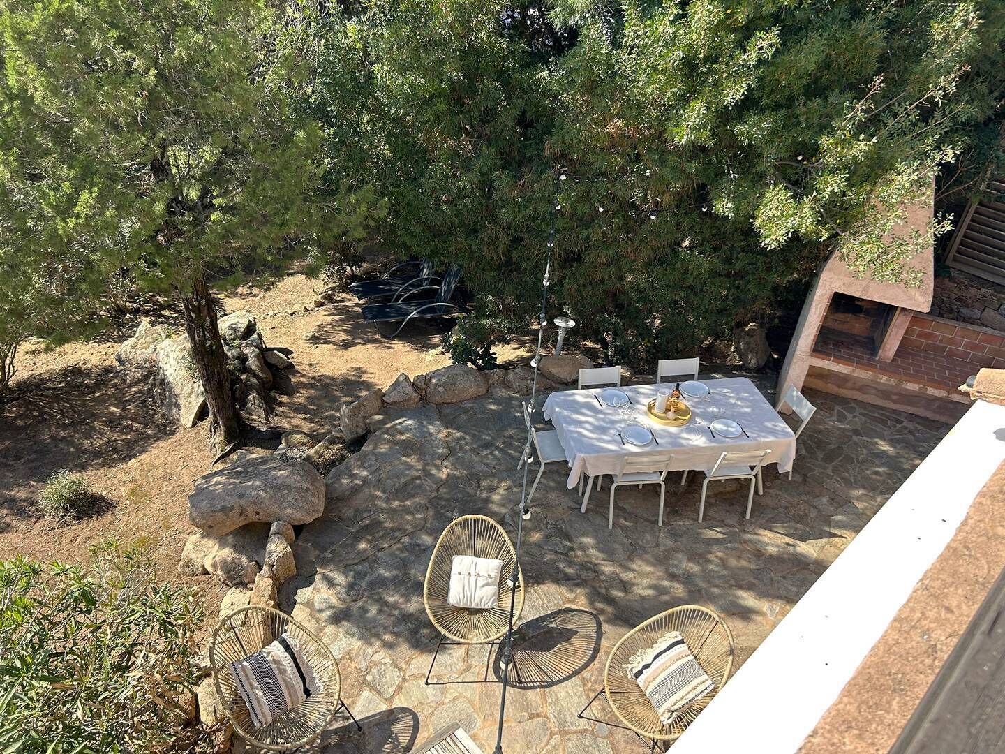 Un'area pranzo all'aperto con un tavolo imbandito per quattro persone, circondata da sedie in vimini, sotto un grande albero.