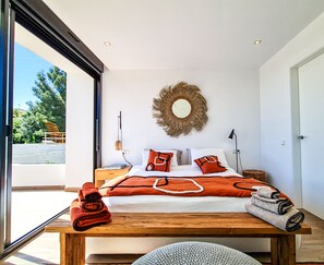 A modern bedroom with a large bed, a wooden nightstand, a round mirror, and a view of the outdoors.