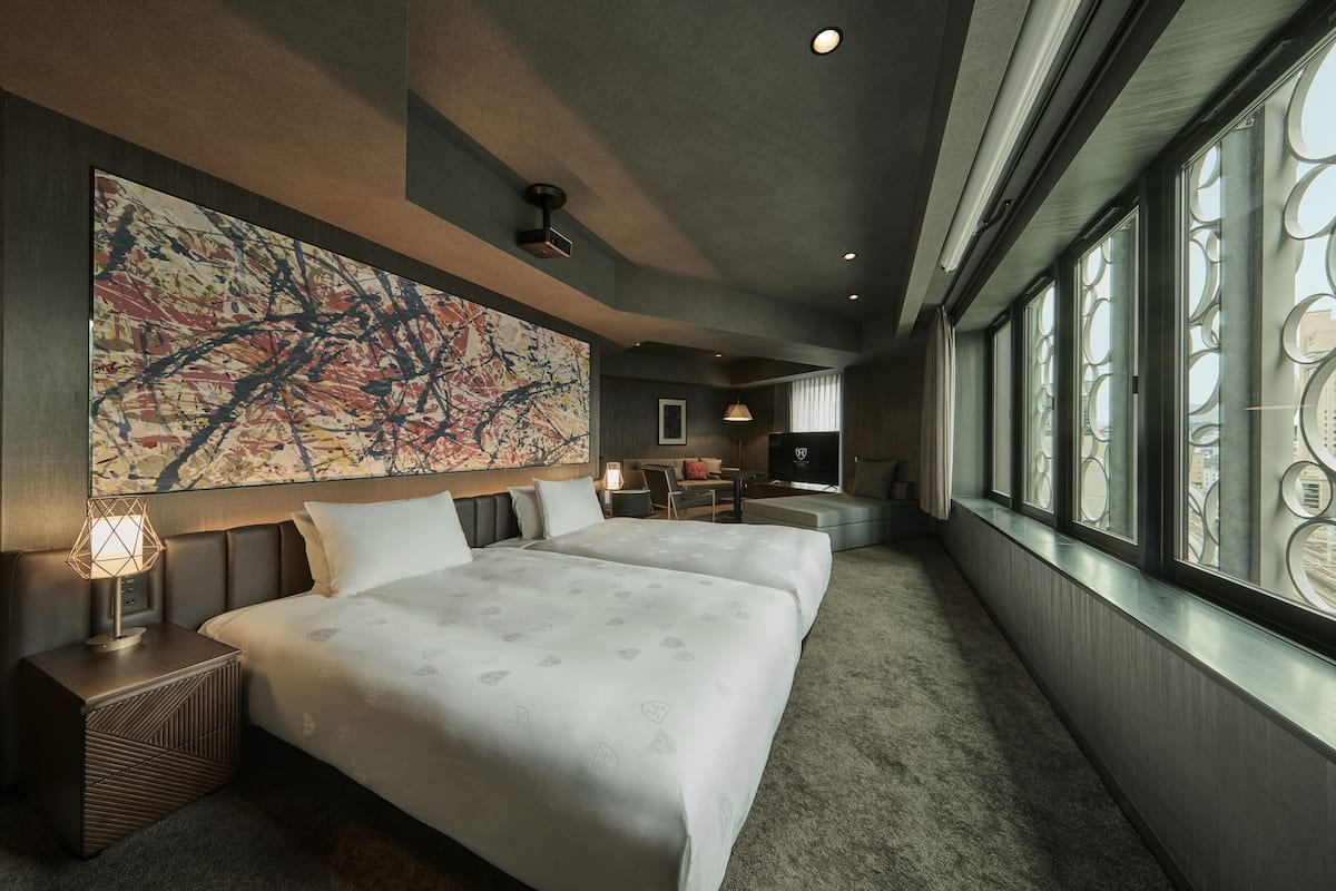 A hotel room with a large abstract painting, two beds, a desk, and a view through circular window frames.
