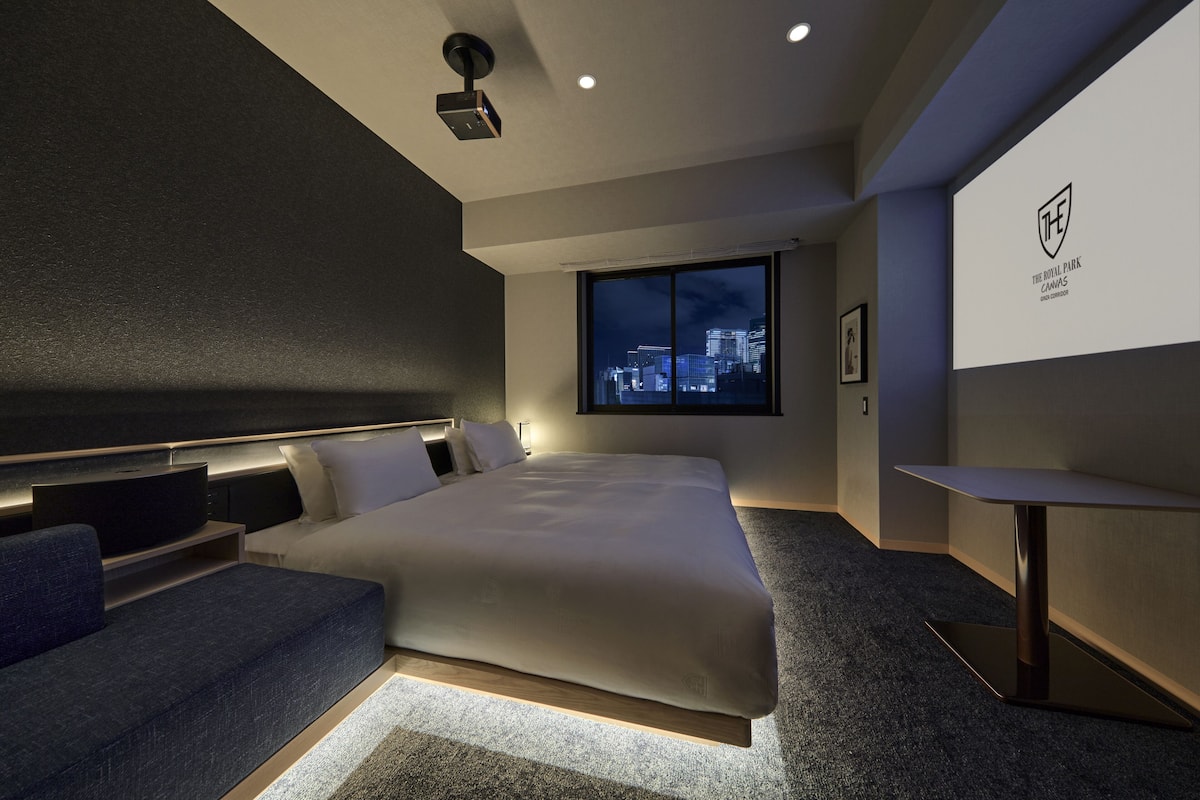 A modern hotel room with a large bed, a seating area, a TV, and a projection screen displaying a logo.