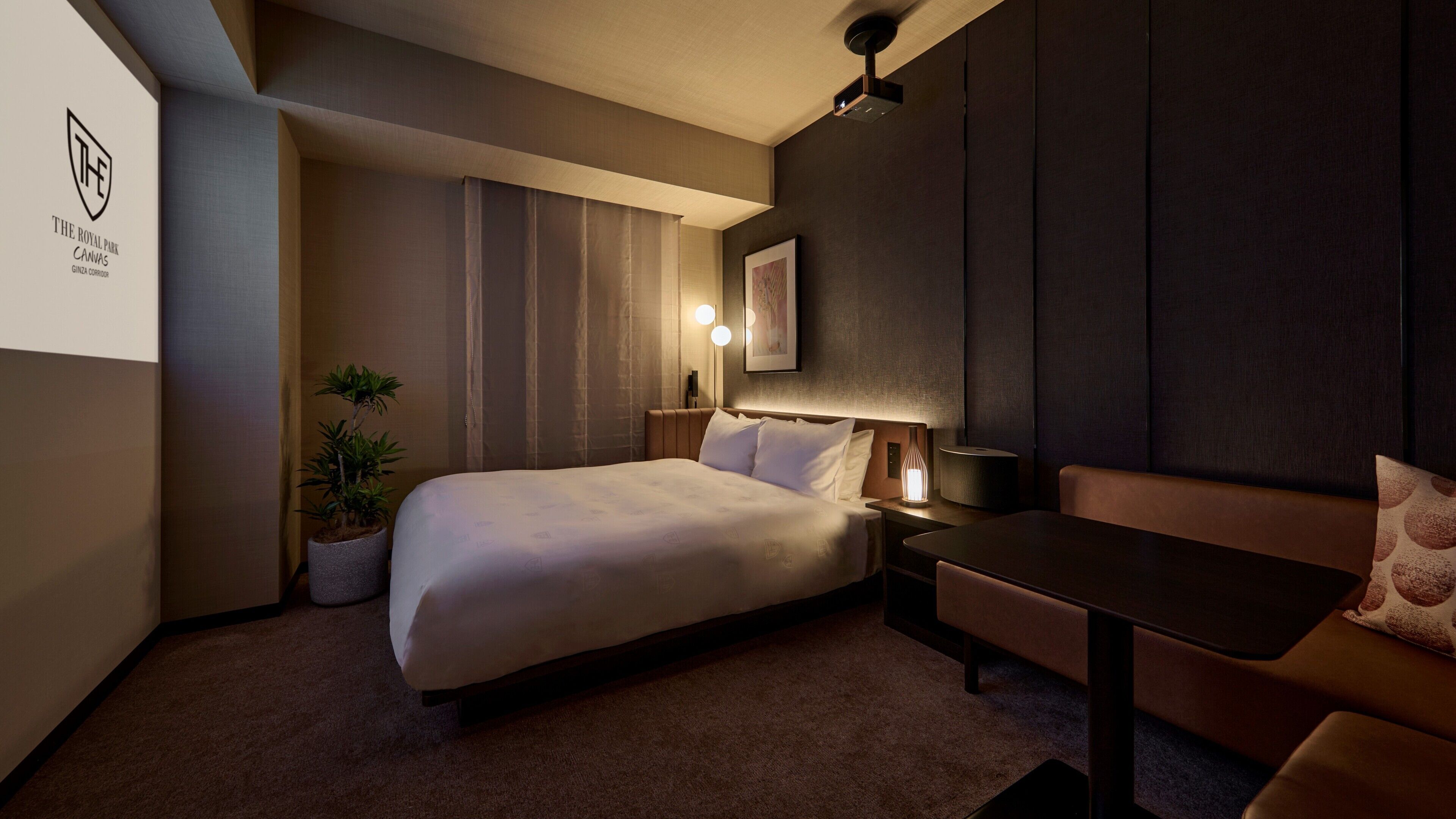Hotelkamer met een bed, bureau, stoel, een potplant en een projector aan de muur.
