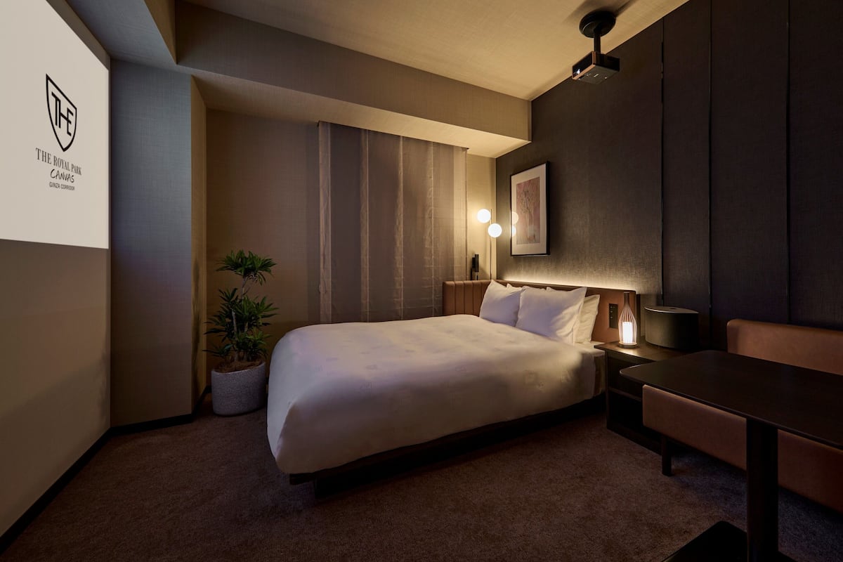 A hotel room with a bed, a desk, a chair, a potted plant, and a wall-mounted projector.