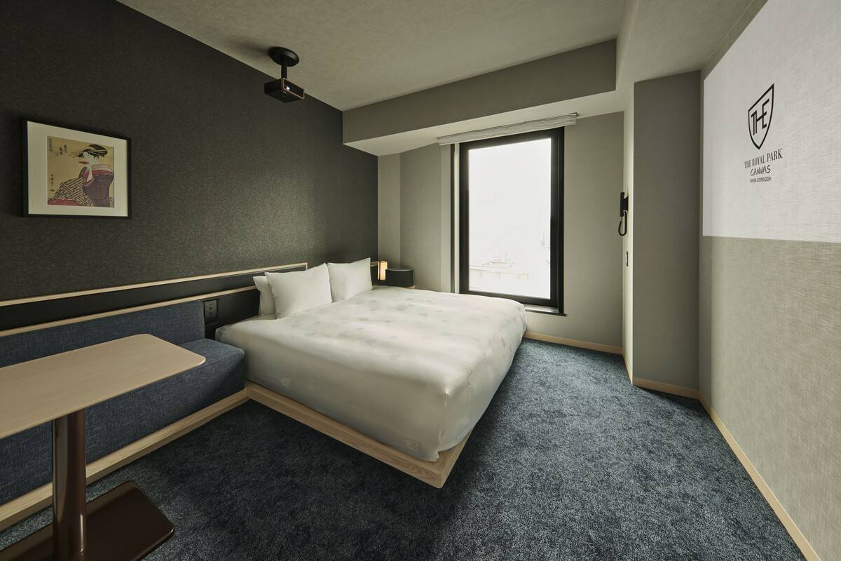 A modern hotel room with a bed, a small desk, a sofa, a window, and a wall-mounted artwork.