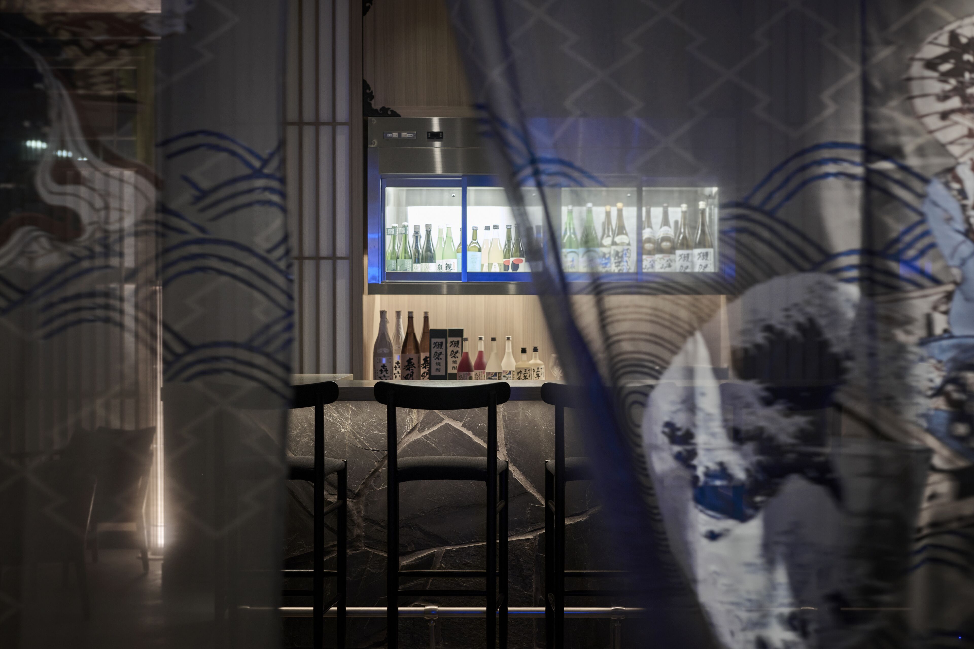 A bar area with a refrigerator stocked with bottles, two black chairs, and a stone wall.