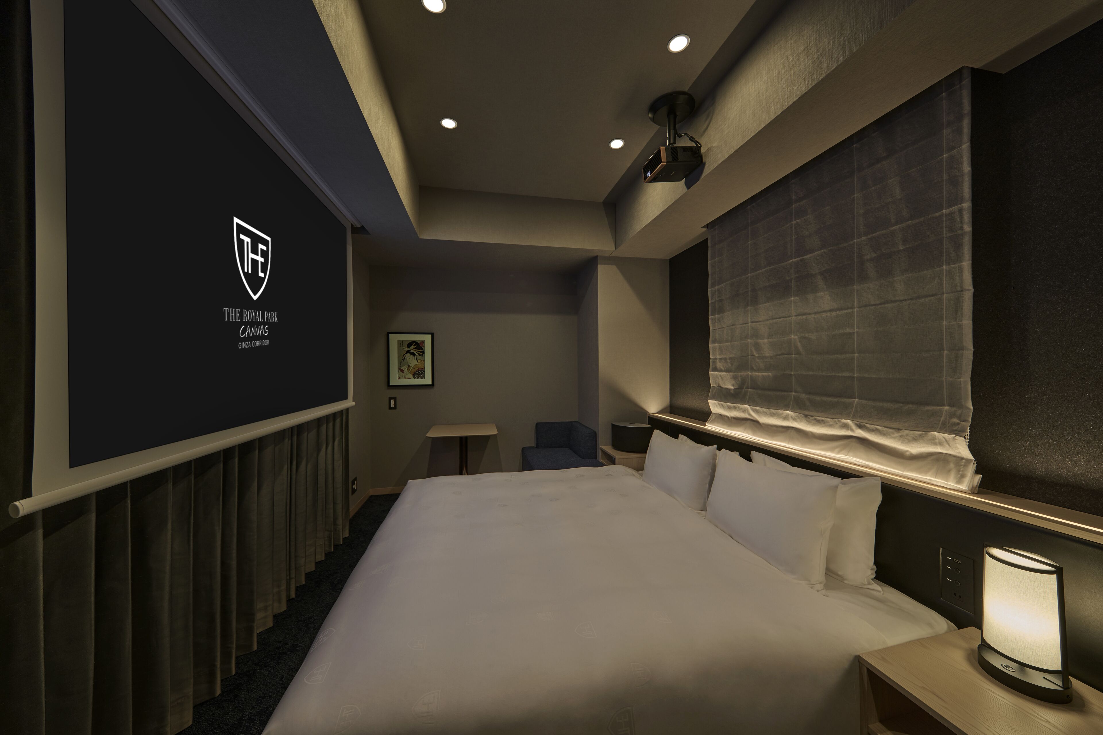 A hotel room with a large bed, a bedside table with a lamp, a small desk, a chair, a projector screen, and a window with curtains.