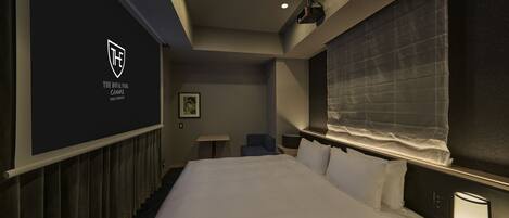 A hotel room with a large bed, a bedside table with a lamp, a small desk, a chair, a projector screen, and a window with curtains.