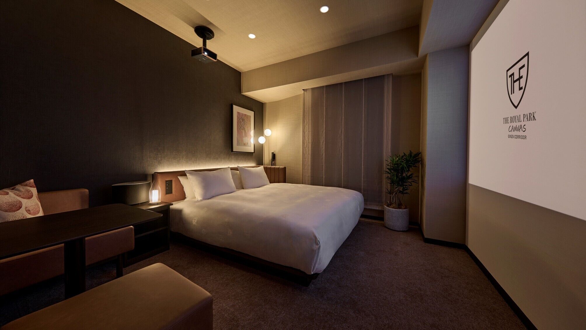 Een hotelkamer met een bed, nachtkastjes, een bureau, een stoel, een kamerplant en een projector bevestigd aan de muur.