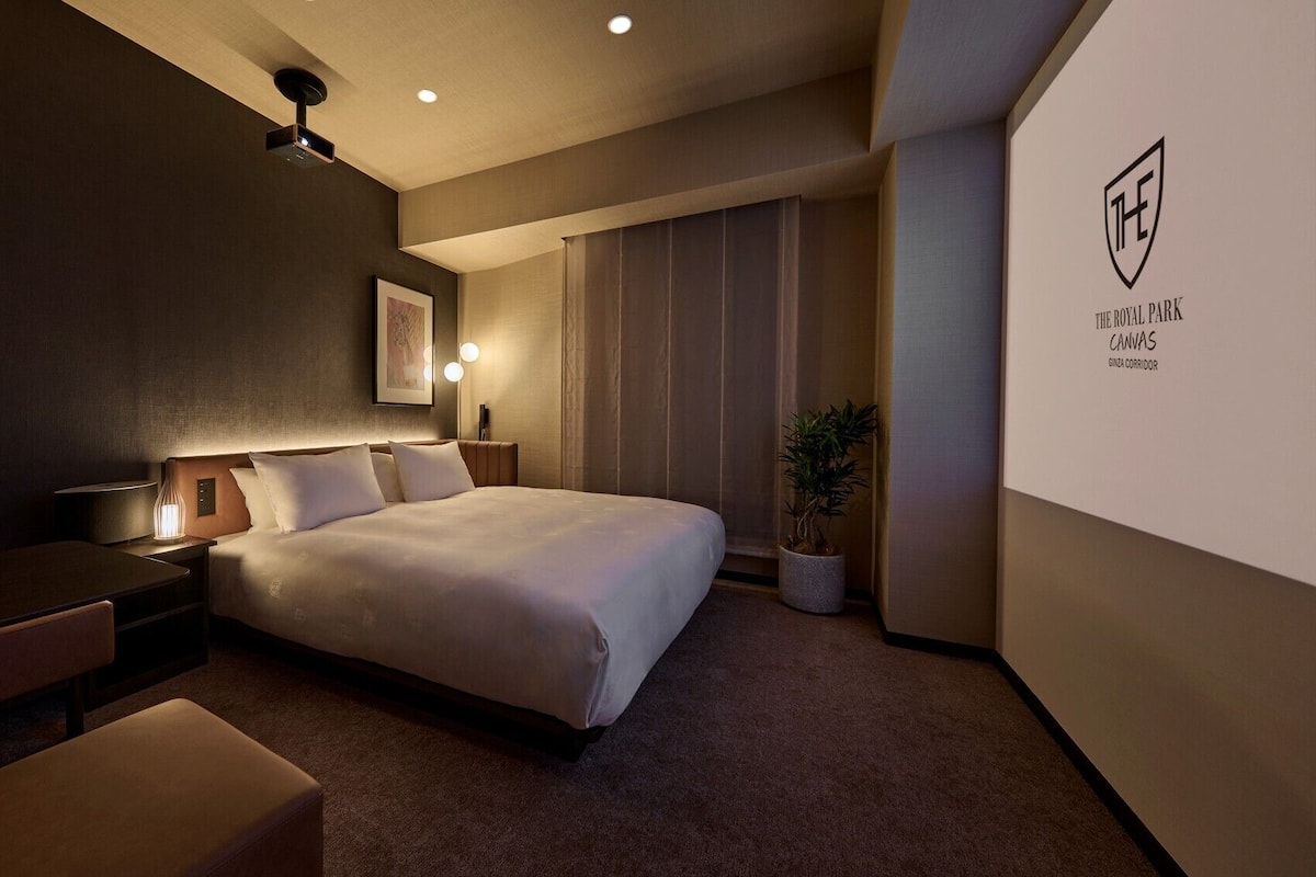 Habitación de hotel con cama, mesitas de noche, escritorio, silla, planta en maceta y proyector fijado a la pared.