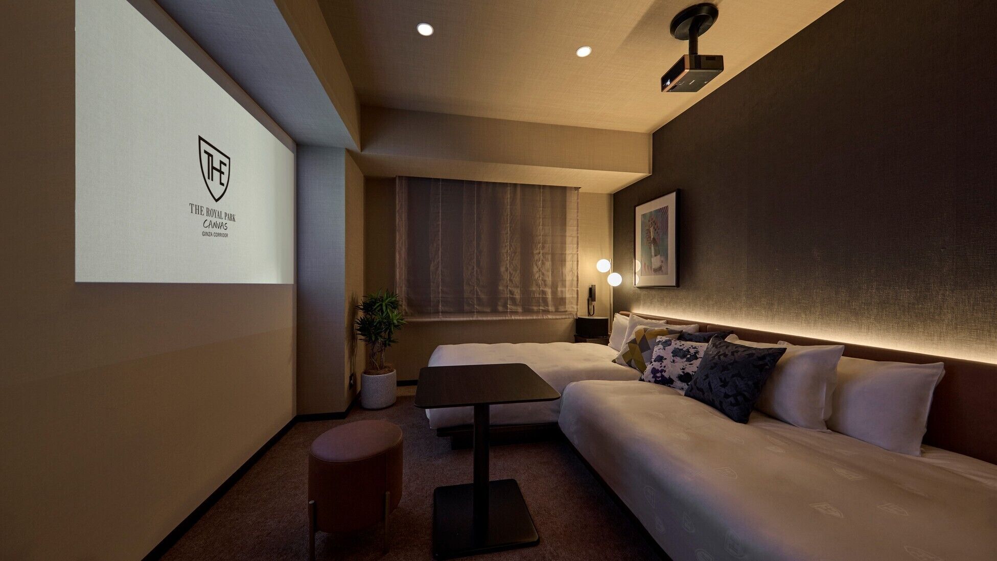 Hotelkamer met een bed, een tafeltje, een scherm voor een projecteerder, een plant en een schilderij aan de muur.