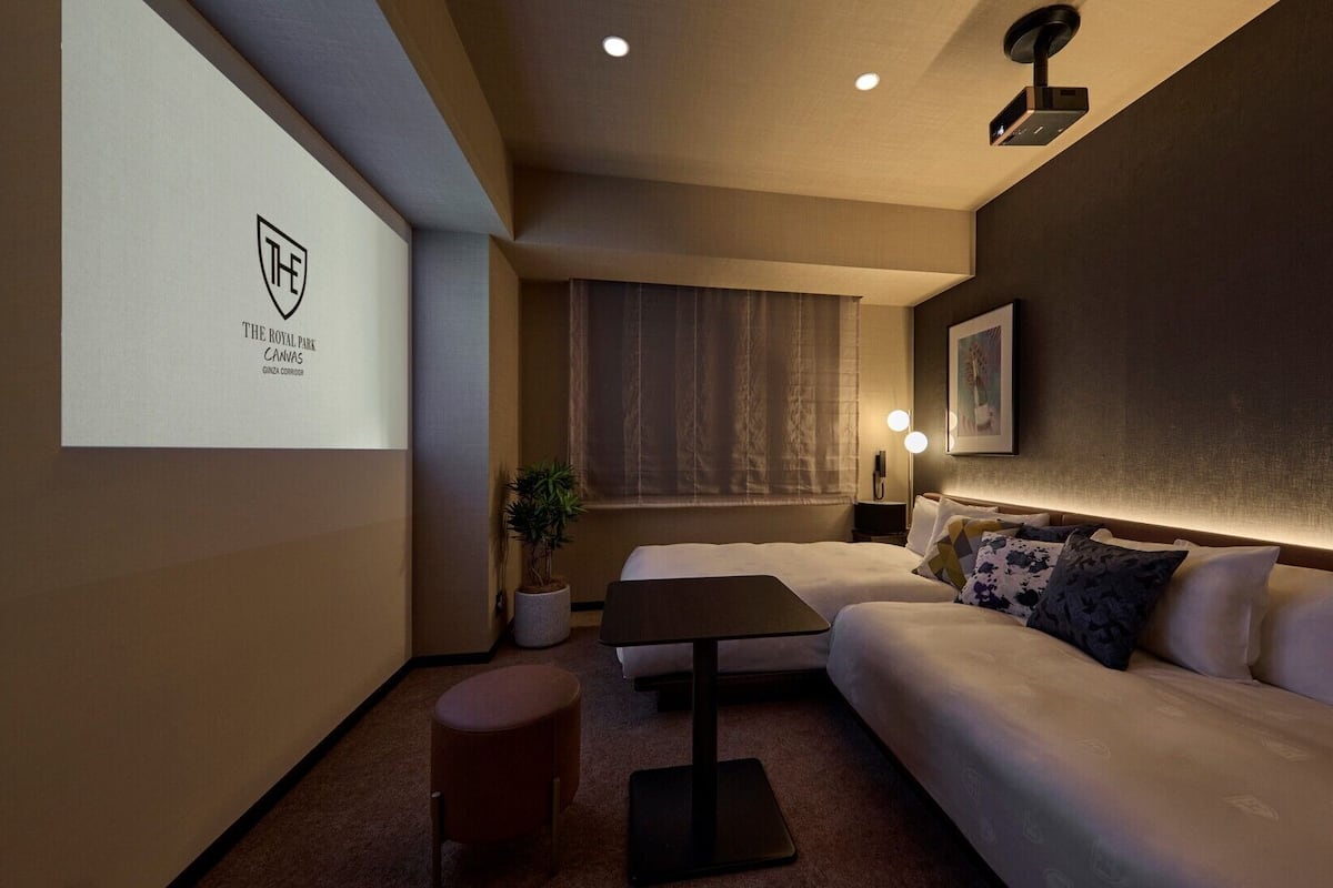 Habitación de hotel con cama, una mesita, pantalla para proyector, una planta y un cuadro en la pared.