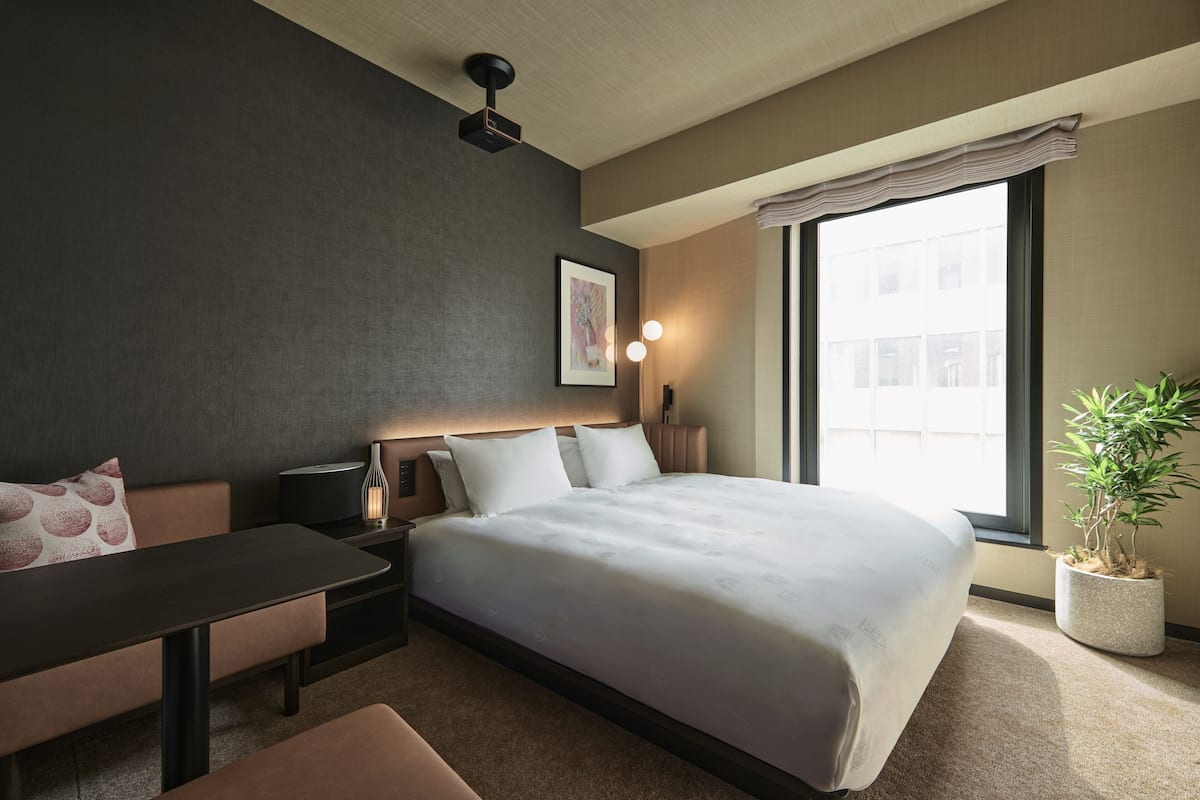A hotel room with a bed, a desk, a chair, a potted plant, and a window with a view of buildings.