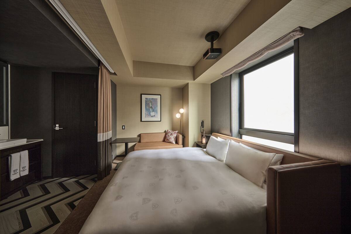 A hotel room with a large bed, a desk, a chair, a nightstand, a lamp, and a window.