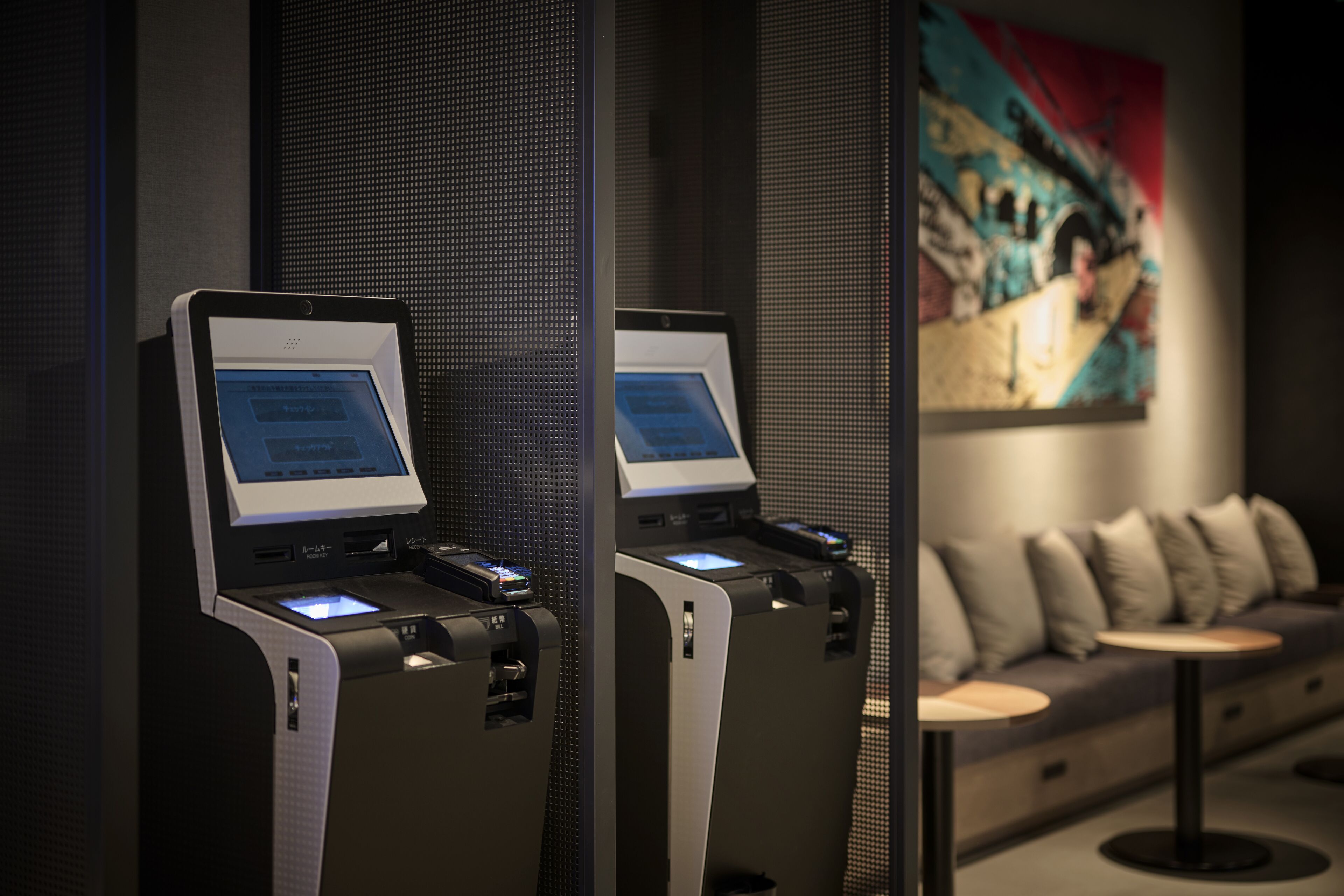 Two self-service kiosks in a modern lobby with a sofa and a colorful painting in the background.