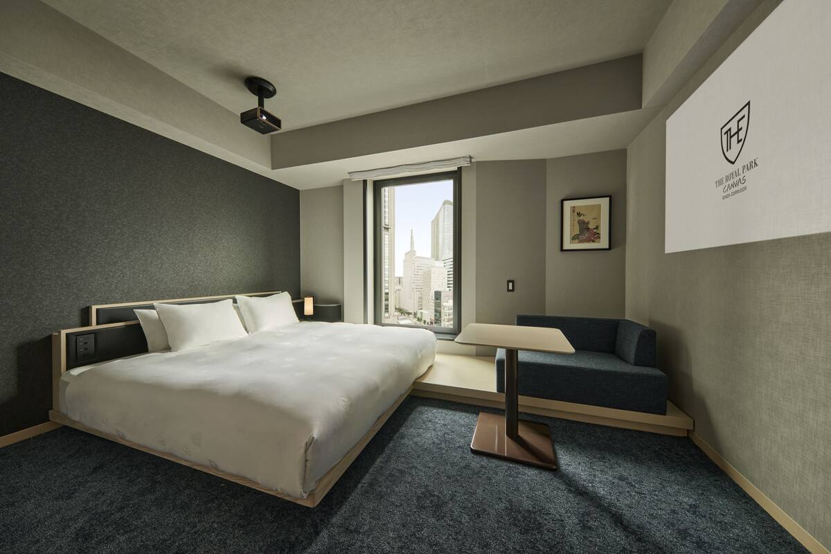 A modern hotel room with a large bed, a seating area, a window with a city view, and a logo on the wall.