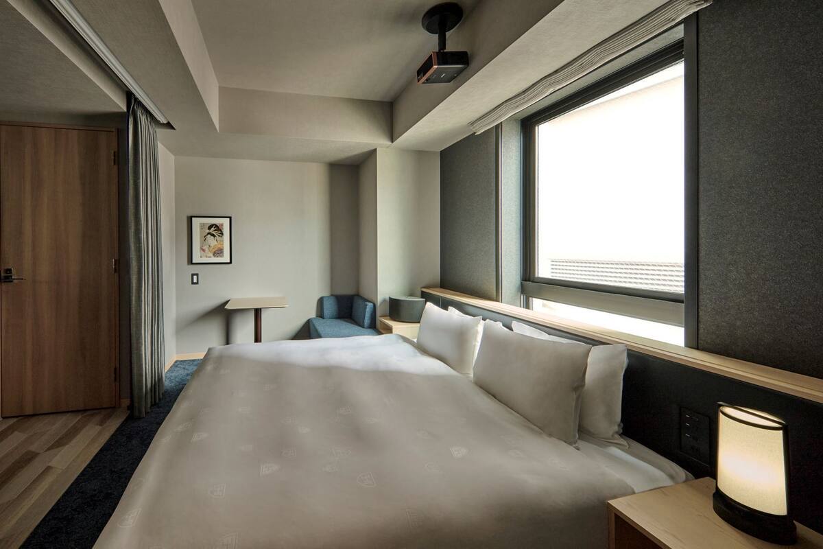 A hotel room with a bed, a desk, a chair, a lamp, and a window.