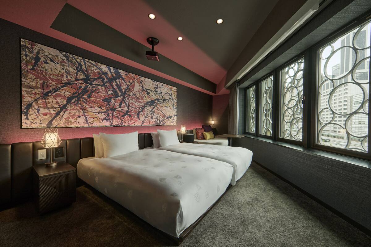 A modern hotel room with a large abstract painting, two beds, a nightstand, and a view of the city through circular window designs.