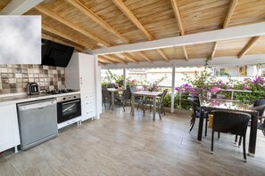 A modern kitchen with a wooden ceiling, a dining area with outdoor seating, and a view of a garden with purple flowers. - Cesme Flowers Butik Otel (Cesme)