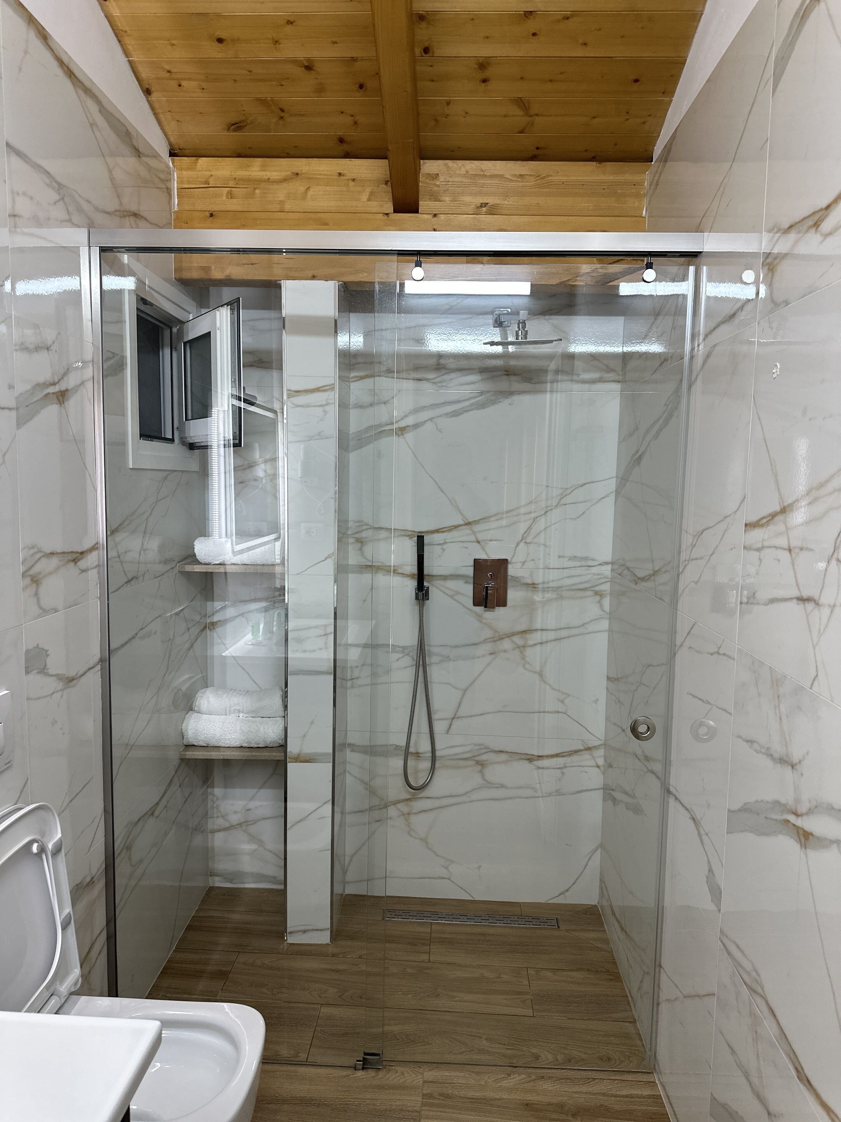 A modern bathroom with a glass-enclosed shower, marble walls, and wooden flooring.
