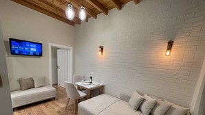 Ein moderner, gut beleuchteter Raum mit weißer Ziegelwand, Holzboden und einem flachbildfernseher an der Wand. Es gibt ein Sofa, einen Esstisch mit Stühlen und einen kleinen Tisch mit Flasche und Gläsern.