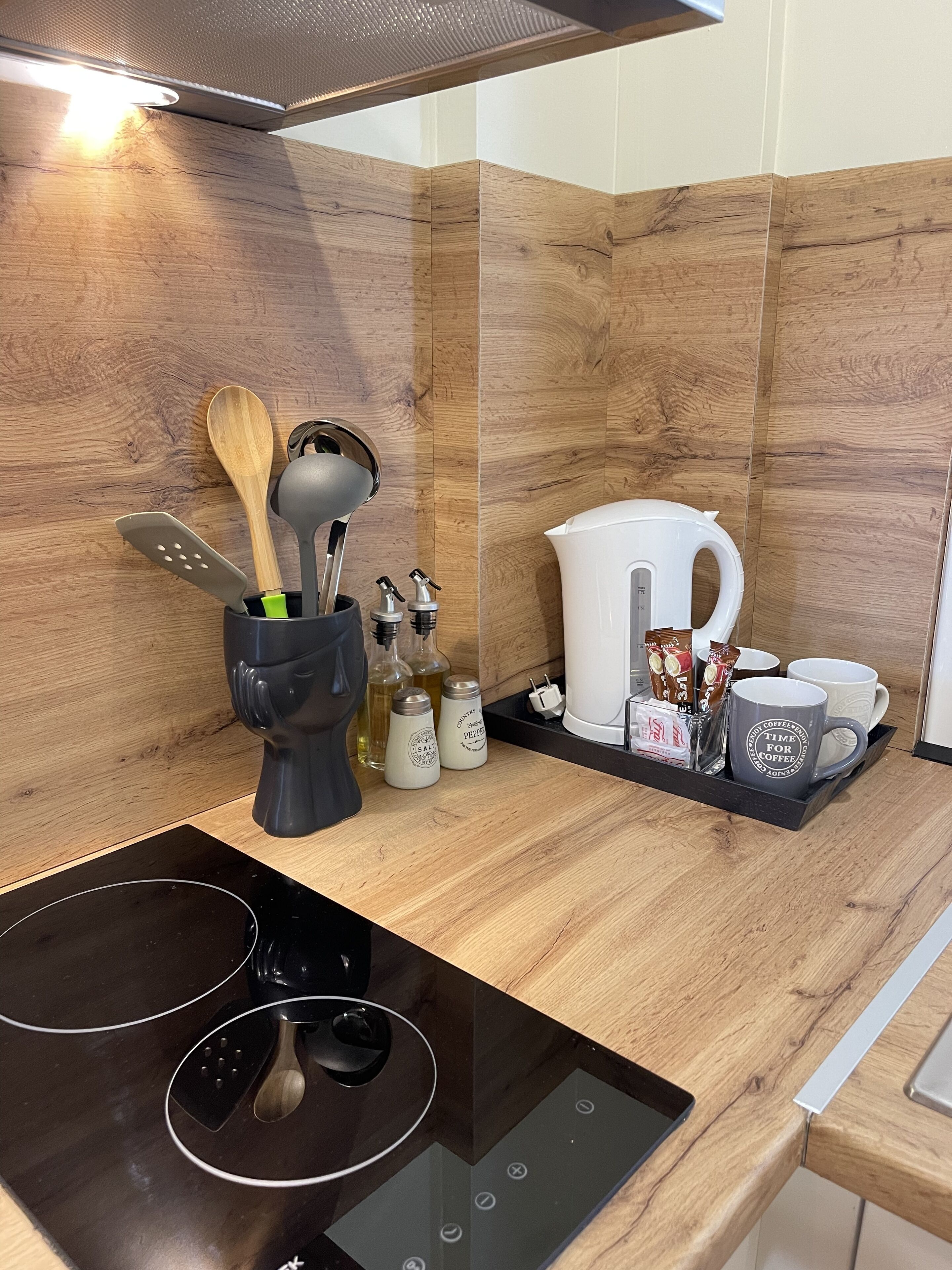 A modern kitchen with a black induction cooktop, wooden backsplash, and a coffee station with mugs and a kettle.
