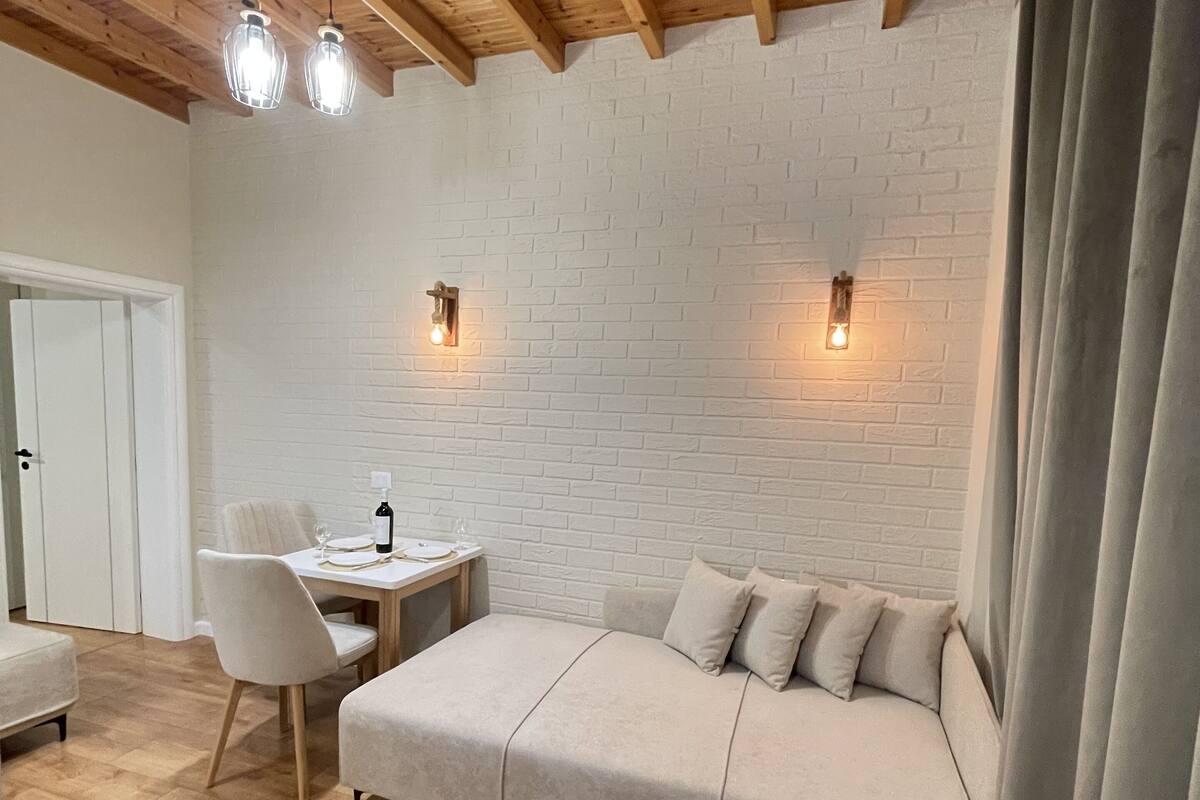 Ein gemütliches Zimmer mit Holzboden, einer weißen Ziegelwand, einer Couch, einem Esstisch mit Stühlen und Pendelleuchten.