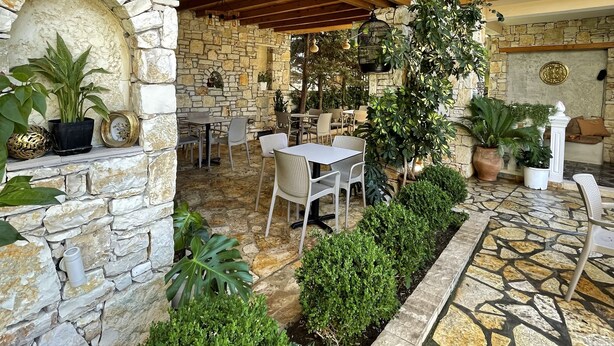 Eine Terrasse im Freien mit Steinsäulen, Tischen und Stühlen, umgeben von Grünpflanzen und einem gemusterten Steinboden.