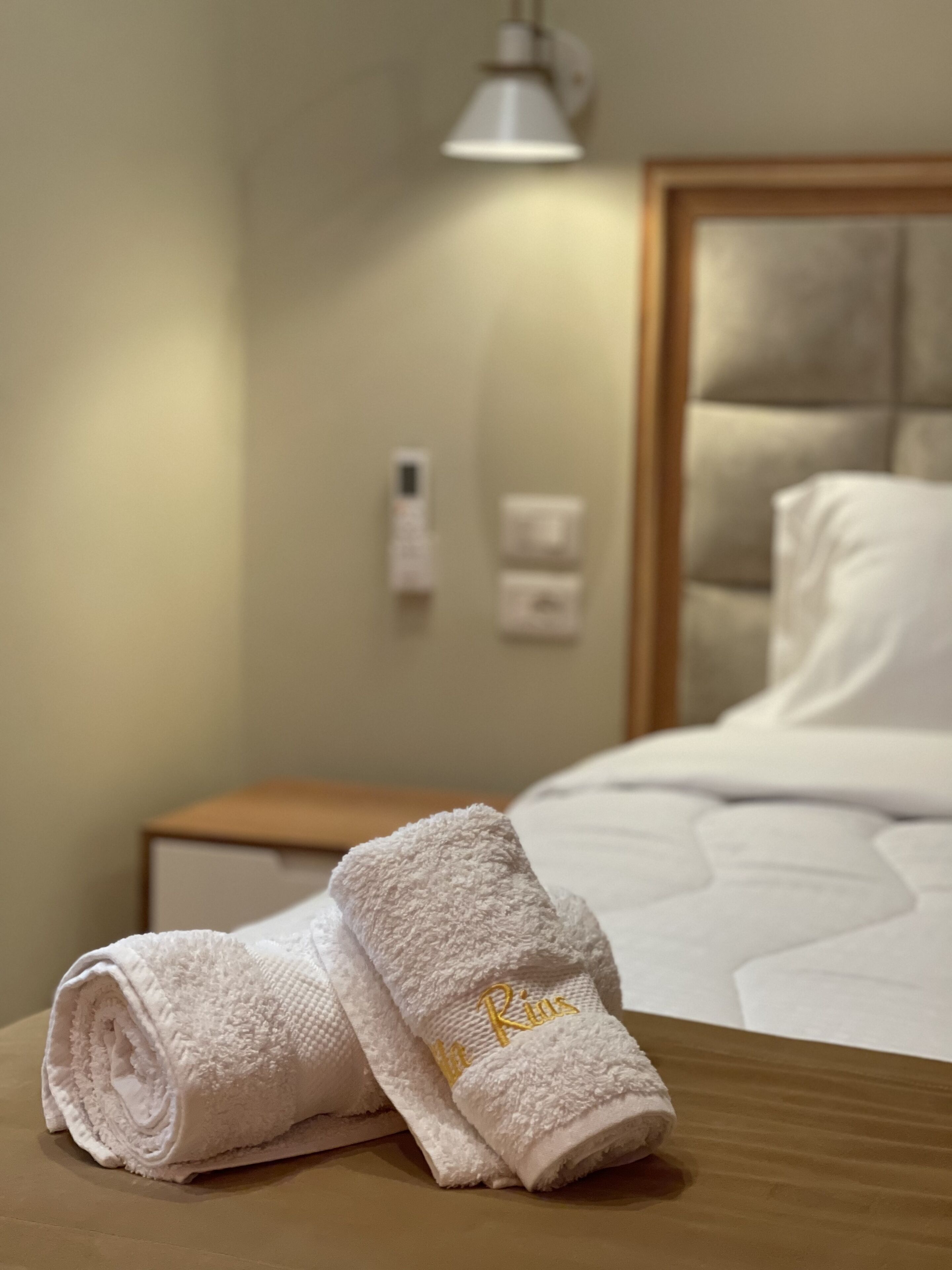 A neatly made bed with a white duvet, a wooden nightstand with a lamp, and two rolled towels with "La Rios" embroidered on them.