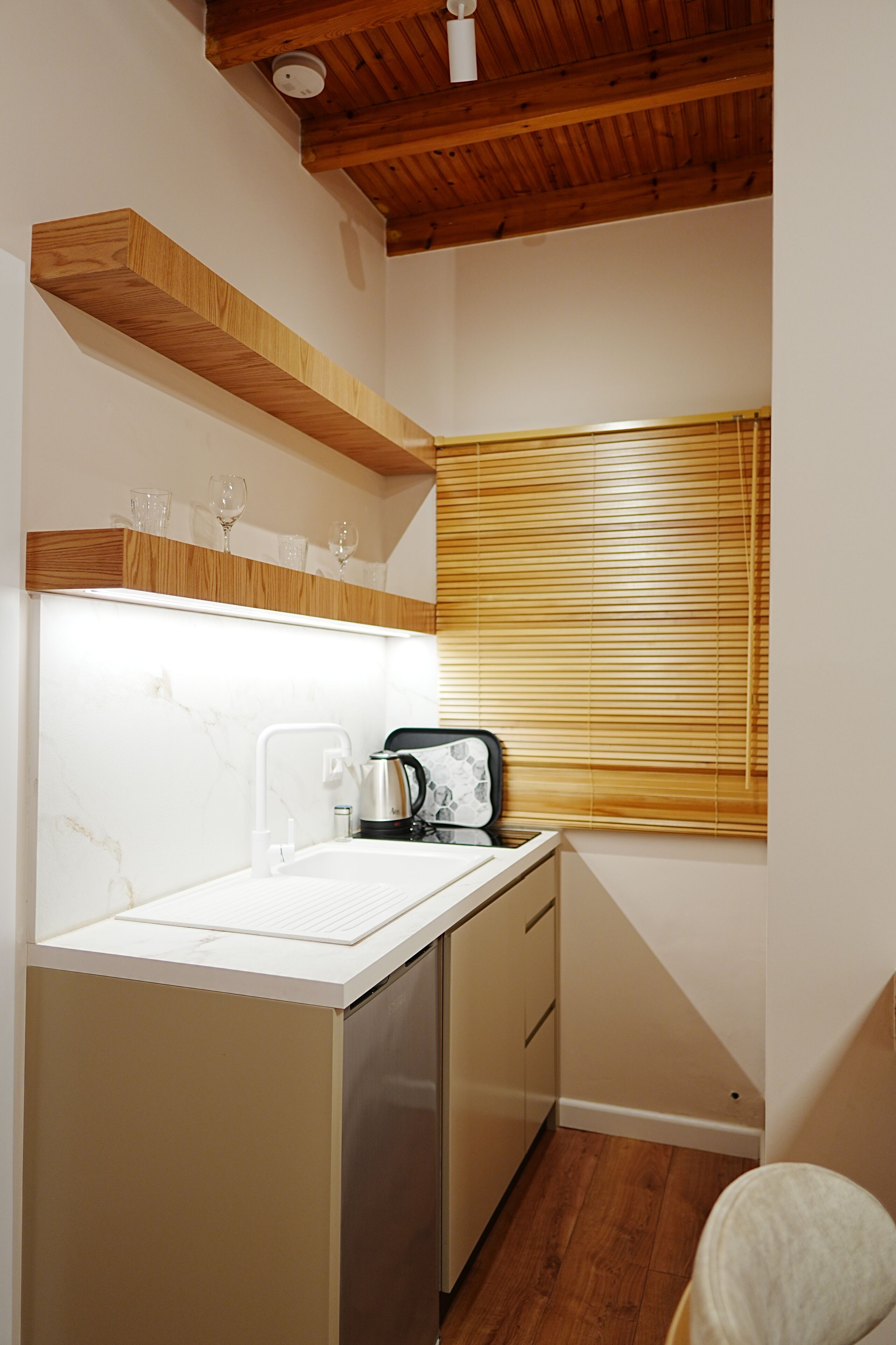 Een kleine keuken met een houten plafond, een wit aanrecht, een inbouwoven en een raam met jaloezieën.